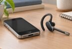 A generic single-ear calling headset beside an older-style smartphone on a desk.