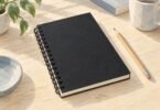 Illustration of a generic closed black wire-bound paper planner beside a pencil.