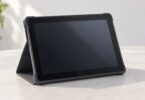 A generic wide-bezel tablet with a blank screen stands in a charcoal folding folio case on a pale tabletop.