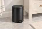 Illustration of a compact matte black pedal bin with a closed lid beside a bathroom vanity.