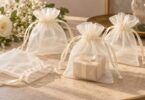 Ivory sheer organza favor pouches with satin drawstrings on a warm neutral table, including one holding a small gift box.