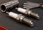 Two automotive spark plugs on a workshop mat with a spark-plug socket and feeler gauge behind them.