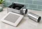 Bathroom exhaust fan housing, white grille and separate short round metal duct on a light surface.
