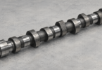 Illustration of a single uninstalled steel camshaft resting on a plain gray tabletop.