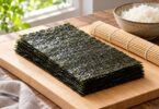 Plain nori sheets beside a bamboo rolling mat and bowl of rice.
