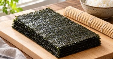 Plain nori sheets beside a bamboo rolling mat and bowl of rice.