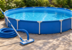 A manual vacuum head, aluminum pole and coiled blue hose on dry paving beside a round blue above-ground pool.