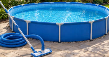 A manual vacuum head, aluminum pole and coiled blue hose on dry paving beside a round blue above-ground pool.