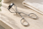 A generic silver eyelash curler with a black pad on a pale stone vanity beside a folded ivory cloth
