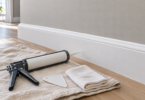 A white baseboard against a light wall, with an unbranded caulk gun, smoothing tool and folded cloth on a drop cloth.