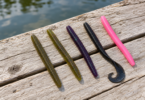 Five soft-plastic fishing worms in green, purple, black and pink lie on a wooden dock beside water.