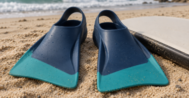 A pair of blue and teal rubber swim fins beside a bodyboard on beach sand.