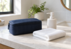 A closed navy fabric pouch, folded white towel and plain white soap dispenser on a bathroom counter.