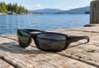 Black wraparound sunglasses with dark lenses resting on a wooden dock beside a lake