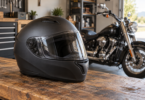 An unbranded matte black full-face helmet with a clear shield on a wooden workbench, with a cruiser motorcycle in the garage behind it.