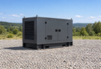 Generic dark gray enclosed generator on a dry gravel clearing with distant trees and blue sky.