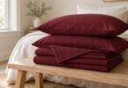 Burgundy pillows and folded sheets stacked on a pale wooden bench in a neutral bedroom.