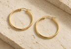 Two gold-colored hoop earrings with open hinged posts resting on a pale stone surface.