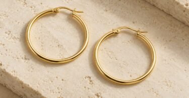 Two gold-colored hoop earrings with open hinged posts resting on a pale stone surface.