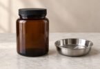 An unlabeled amber glass jar with a closed black lid beside an empty stainless steel dish on a pale stone counter.