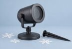 Illustration of an unbranded dark projector on a round base beside a garden stake and paper snowflakes