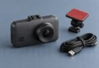 Generic dark dashboard camera with a detached adhesive mount and coiled USB cable on a blue-gray surface.