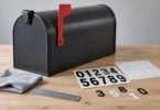 Blank charcoal mailbox beside number decal sheets, loose digits, a ruler and a cloth on a tabletop