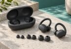 Generic dark sport earbuds with ear hooks, an open charging case and three pairs of silicone tips on a stone surface.