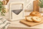 Illustration of a generic guarded countertop bagel slicer beside a whole sesame bagel and two halves.
