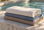 Ivory, slate-blue and taupe towels folded on a stone ledge beside blue spa water.