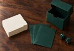 Illustrative still life of blank trading cards, green sleeves, a deck box and dice on a wooden table.