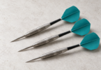 Illustration of three generic steel-tip darts with silver barrels, black shafts and teal flights on a pale tabletop