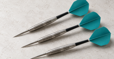 Illustration of three generic steel-tip darts with silver barrels, black shafts and teal flights on a pale tabletop