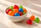 Generic red, orange, green and blue soft candies in a white bowl with three loose candies on a light tabletop.