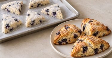Unbaked blueberry scone portions on a parchment-lined tray beside golden baked scones on a plate