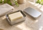 A plain soap bar on a perforated tray inside an open silver metal tin, with its lid and a folded washcloth beside it.
