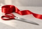 Illustration of closed red-handled silver-colored scissors resting on a neutral table beside a roll of red satin ribbon