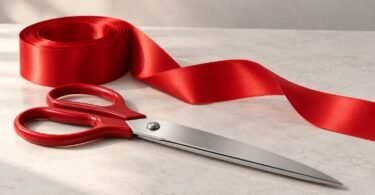 Illustration of closed red-handled silver-colored scissors resting on a neutral table beside a roll of red satin ribbon