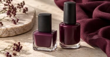 Two unbranded bottles of plum and wine-colored nail polish on pale stone beside burgundy fabric
