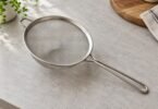 Unbranded stainless-steel mesh strainer with a long loop handle and an opposing rest on a light kitchen counter.