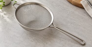 Unbranded stainless-steel mesh strainer with a long loop handle and an opposing rest on a light kitchen counter.