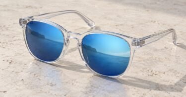 A pair of transparent-frame sunglasses with blue mirrored lenses resting on a pale stone tabletop.