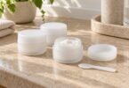 Three plain white cream jars, one open beside its lid and a small cosmetic spatula on a stone counter.