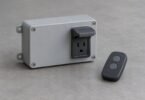Unbranded enclosed electrical control device with an empty outlet beside a two-button remote on a gray workbench