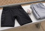 Generic black padded sports shorts beside a baseball and folded gray uniform pants on a bench