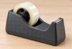 A dark desktop tape dispenser holding clear tape on a pale wood table.