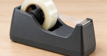A dark desktop tape dispenser holding clear tape on a pale wood table.
