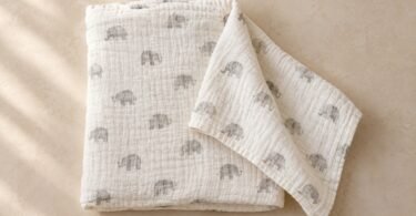 Folded ivory fabric blanket with small gray elephant prints and one corner spread out on a beige surface.