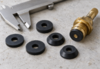 Black flat and beveled rubber washers lie beside a metal caliper and a brass faucet stem.