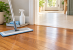 A flat microfiber mop on wood flooring beside a white spray bottle and folded cloth, with a tiled doorway behind.